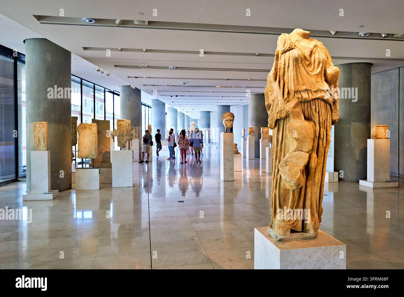Besucher gehen an einer kopflosen, drapierten Marmorstatue und antiken griechischen Reliefs auf Sockeln in einer Galerie mit Betonssäulen vorbei, Griechenland Stockfoto