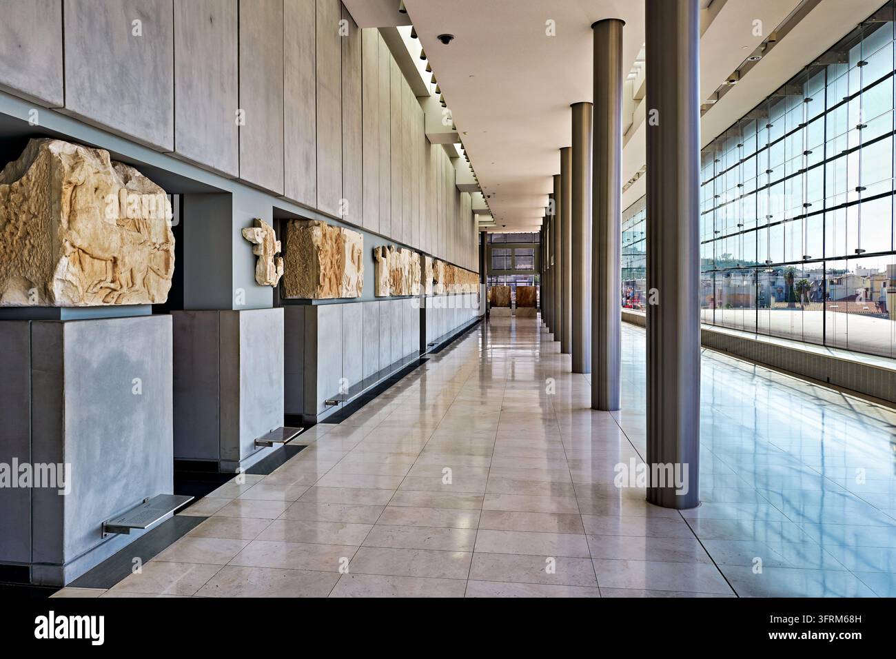 Alte Marmorfriesreliefs säumen einen sonnenbeschienenen Korridor mit Metallssäulen und einer Glaswand mit Blick auf die Stadt, Athen, Griechenland Stockfoto