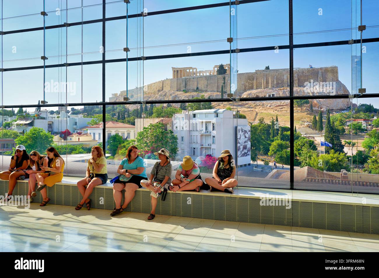 Touristen sitzen neben einer bodentiefen Glaswand mit Blick auf den Akropolis-Hügel und den Parthenon-Tempel an einem sonnigen Juninachmittag in Griechenland. Stockfoto