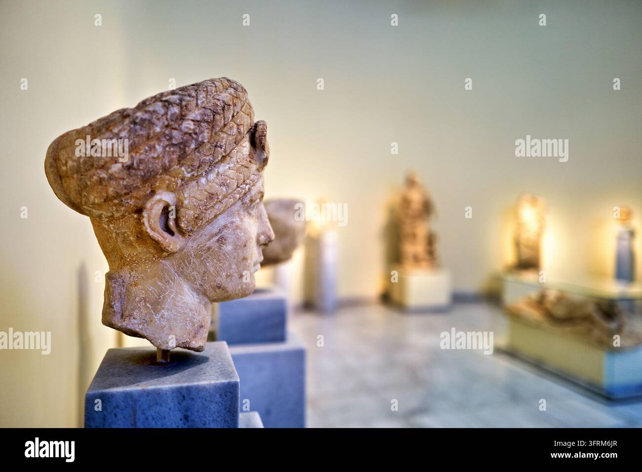 Geformter Kopf mit geflochtener Frisur auf einem Steinpodest in einer Archäologiegalerie, mit verschwommenen Statuen im Hintergrund. Stockfoto