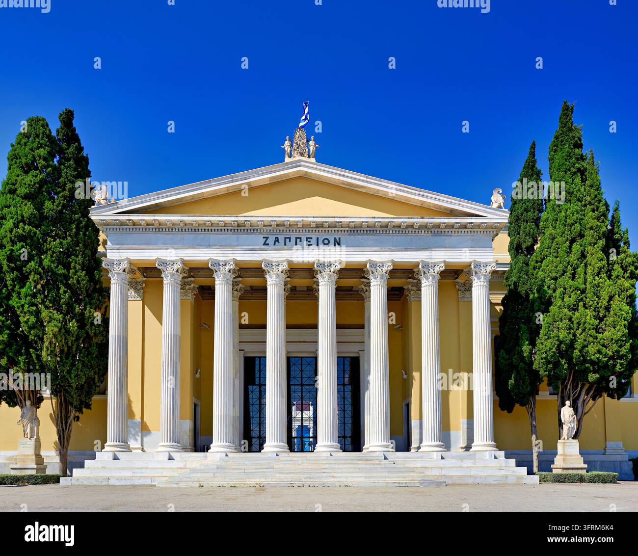 Korinthische Marmorsäulen des Zappeion im Nationalgarten von Athen, mit Zypressen und einer griechischen Flagge vor einem klaren blauen Sommerhimmel Stockfoto
