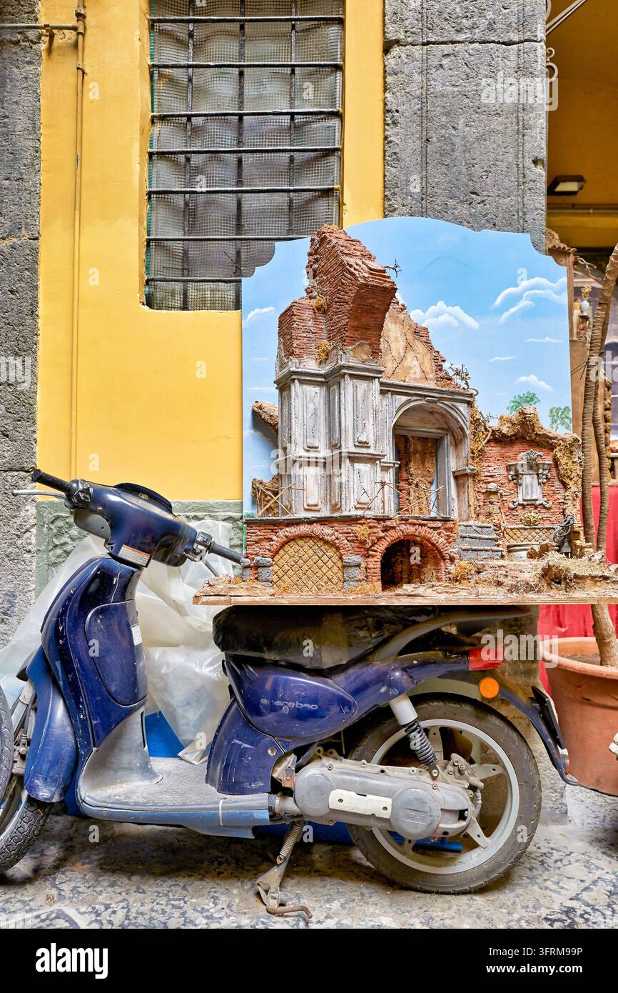 Handgemachtes neapolitanisches Presepe mit Ziegelruinen und gemaltem Himmel ruht auf einem geparkten Motorroller neben einer gelben Mauer in einer alten Stadtstraße, Kampanien Stockfoto