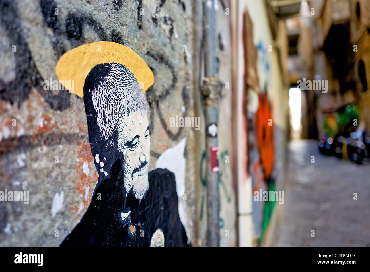 Aufgeklebtes Kunstwerk eines lächelnden bärtigen Mannes mit einem goldenen Heiligenschein auf einer mit Graffiti bedeckten Wand in einer schmalen gepflasterten Gasse in Neapel, Süditalien Stockfoto