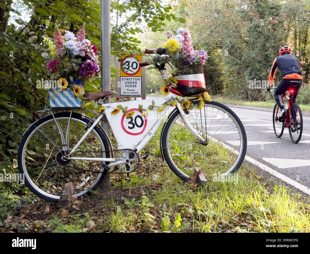 Blumenverziertes Fahrrad neben einem Geschwindigkeitsschild mit einer Geschwindigkeitsbegrenzung von 30 Meilen pro Stunde an der Bezirksgrenze in Stretton unter Fosse, Warwickshire, England. Stockfoto