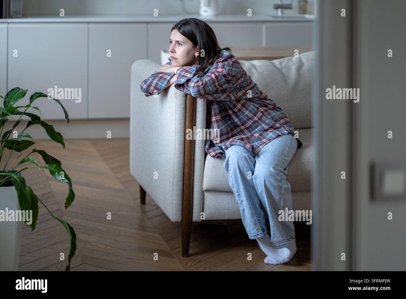 Einsame junge Frau sitzt auf dem Sofa mit leerem Blick, fühlt sich emotional erschöpft, in Gedanken versunken. Stockfoto