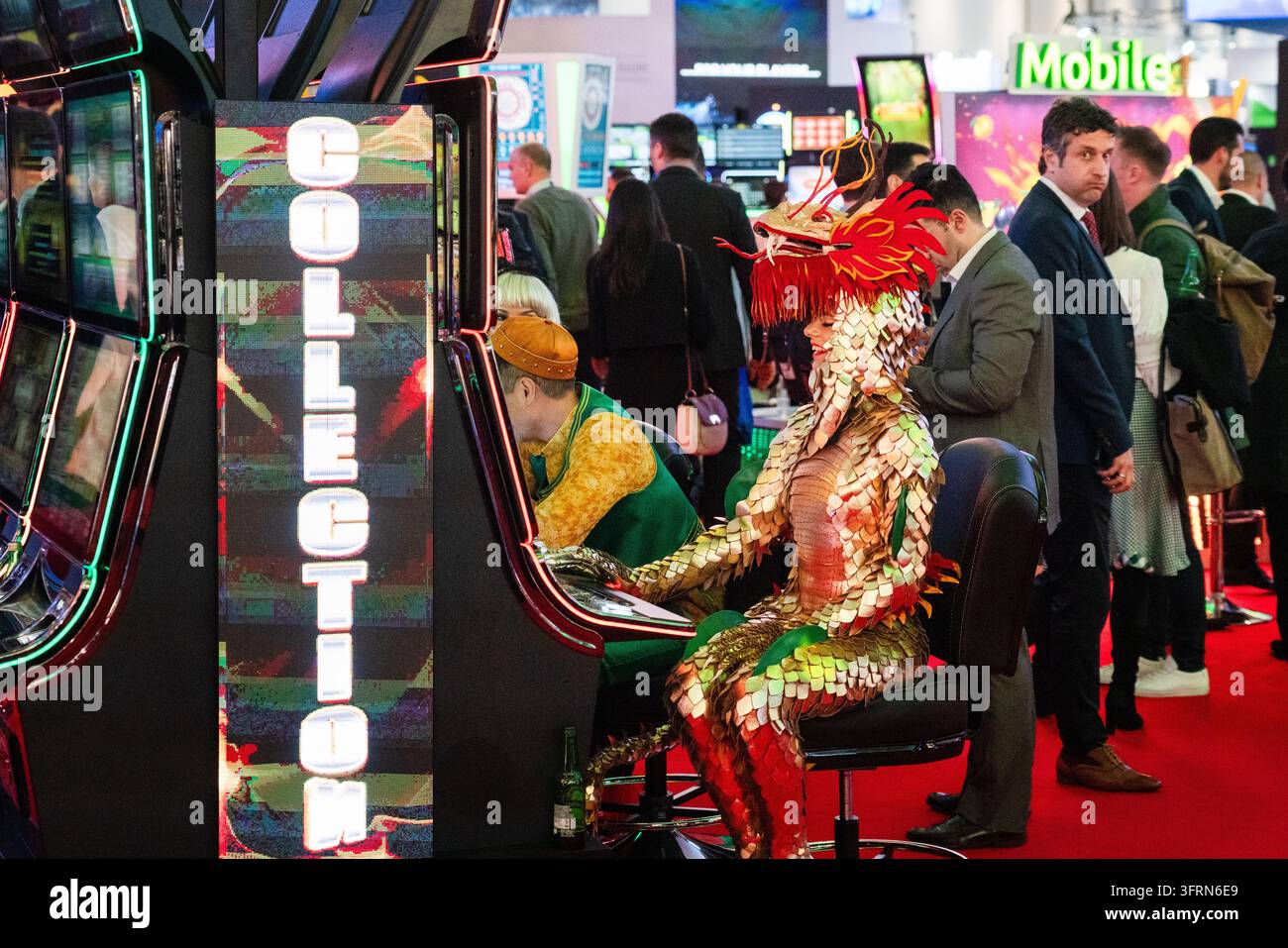 PERSON IM DRACHENKOSTÜM SPIELT EINEN SPIELAUTOMATEN, ICE GAMBLING AND GAMING CONVENTION, LONDON, ENGLAND, 2019: Bei einer lebendigen Kasinoausstellung sitzt eine Person in einem aufwendigen rot-goldenen Drachenkostüm und spielt neben einem Mann in einem gelb-grünen Outfit einen Spielautomaten. INFO: ICE London war historisch die weltweit führende Business-to-Business-Messe für die Glücksspielindustrie, die jährlich im Ausstellungszentrum ExCeL stattfand. Organisiert von Clarion Gaming, verlegte das riesige Ereignis offiziell 2025 nach Barcelona, um seine massive globale Expansion zu ermöglichen. Stockfoto