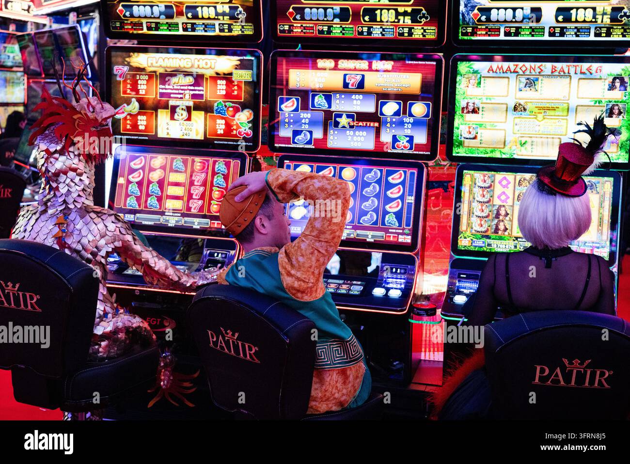 KOSTÜMIERTER SPIELER KRATZT SICH AM SPIELAUTOMAT, ICE GAMBLING UND GAMING KONVENTION, LONDON, ENGLAND, 2019: Ein Mann in einem Volkskostüm kratzt sich in einem Moment der Verwirrung oder Frustration am Kopf, während er einen hell erleuchteten Spielautomaten bedient, zwischen zwei anderen Personen in aufwendigen Kostümen sitzend. INFO: ICE London war historisch die weltweit führende Business-to-Business-Messe für die Gaming- und Glücksspielindustrie, die jährlich im ExCeL-Ausstellungszentrum stattfand. Organisiert von Clarion Gaming, verlegte die riesige Veranstaltung offiziell 2025 nach Barcelona, um ihre massive globale Expansion zu bewältigen. Stockfoto