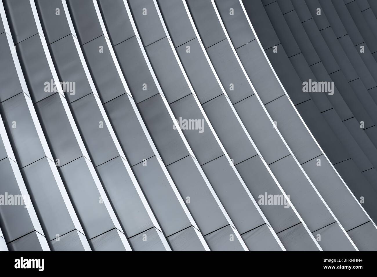 Nahaufnahme einer abstrakten Ansicht einer zeitgenössischen Hochhausfassade mit sich wiederholenden dunkelgrauen Metallpaneelen, hellen linearen Spalten und diagonaler Perspektive, die erschafft Stockfoto