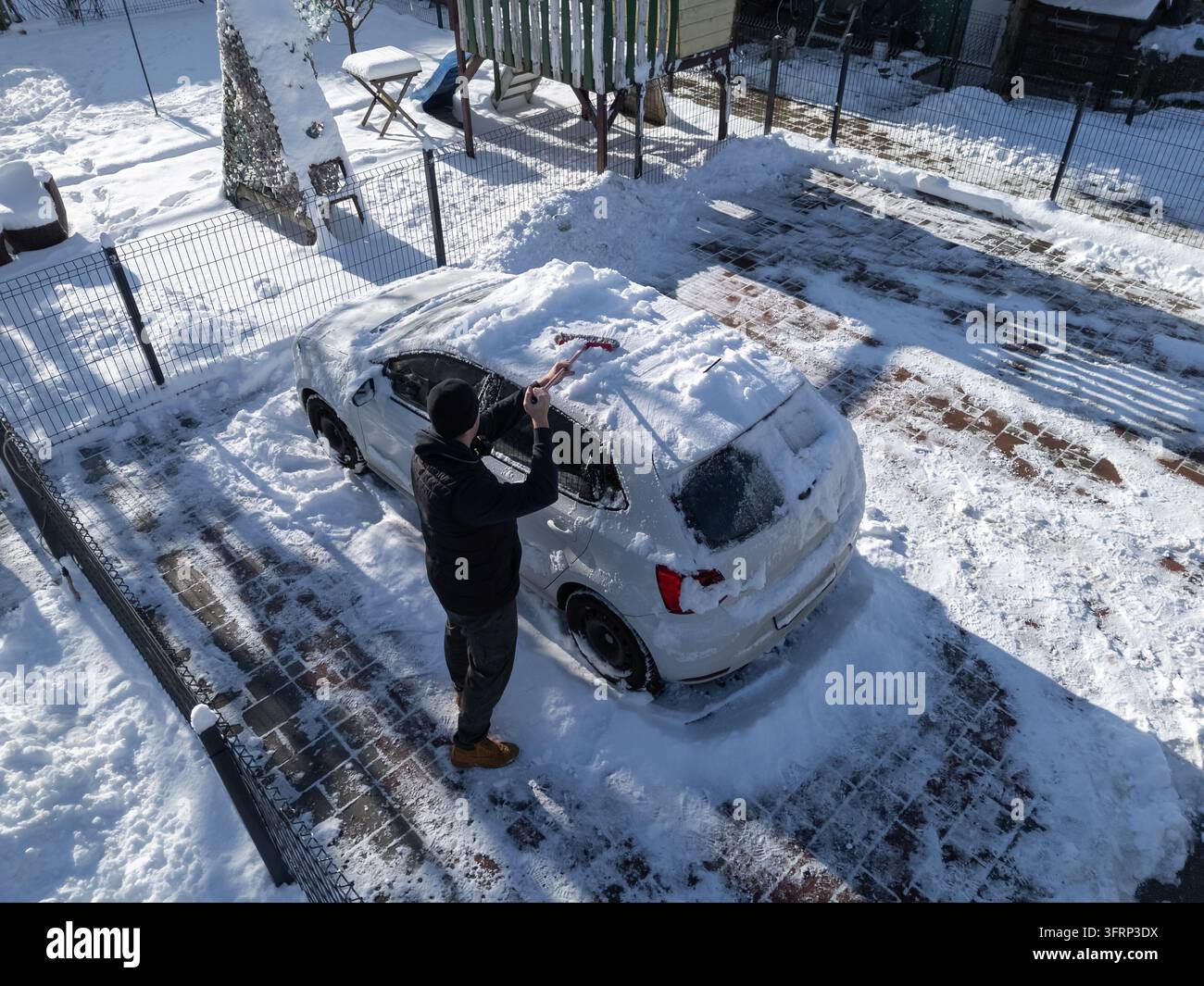 Ein Mann räumt Schnee von einem Auto, das an einem kalten Wintermorgen in einem verschneiten Hof geparkt ist, während er sich auf das Fahren bei Winterwetter vorbereitet. Stockfoto