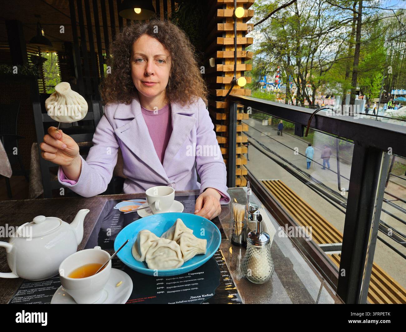 Frau sitzt an einem Tisch mit einem Teller Essen vor sich. Sie hält ein Knödel in der Hand. Text der Speisekarte auf dem Tisch. Stockfoto