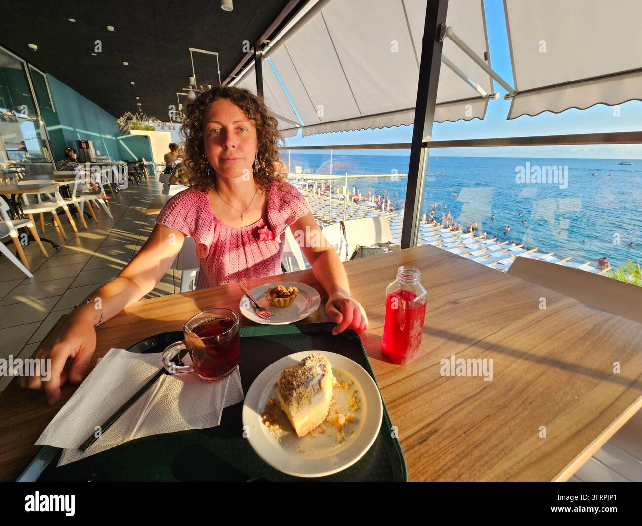 Frau sitzt an einem Tisch mit einem Teller Kuchen und einer Tasse Tee. Sie trägt ein rosa Hemd im Esszimmer im zweiten Stock am Strand in Sotschi Stockfoto