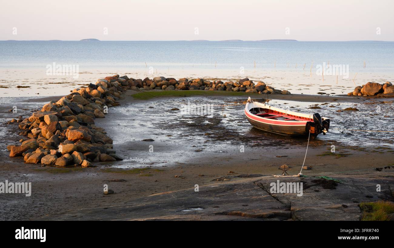 Ein kleines, offenes Motorboot ruht auf einer schlammigen Priel während der Ebbe. Das Boot, weiß, rot und schwarz gestrichen, ist mit einem Seil an das felsige Ufer befestigt. Stockfoto
