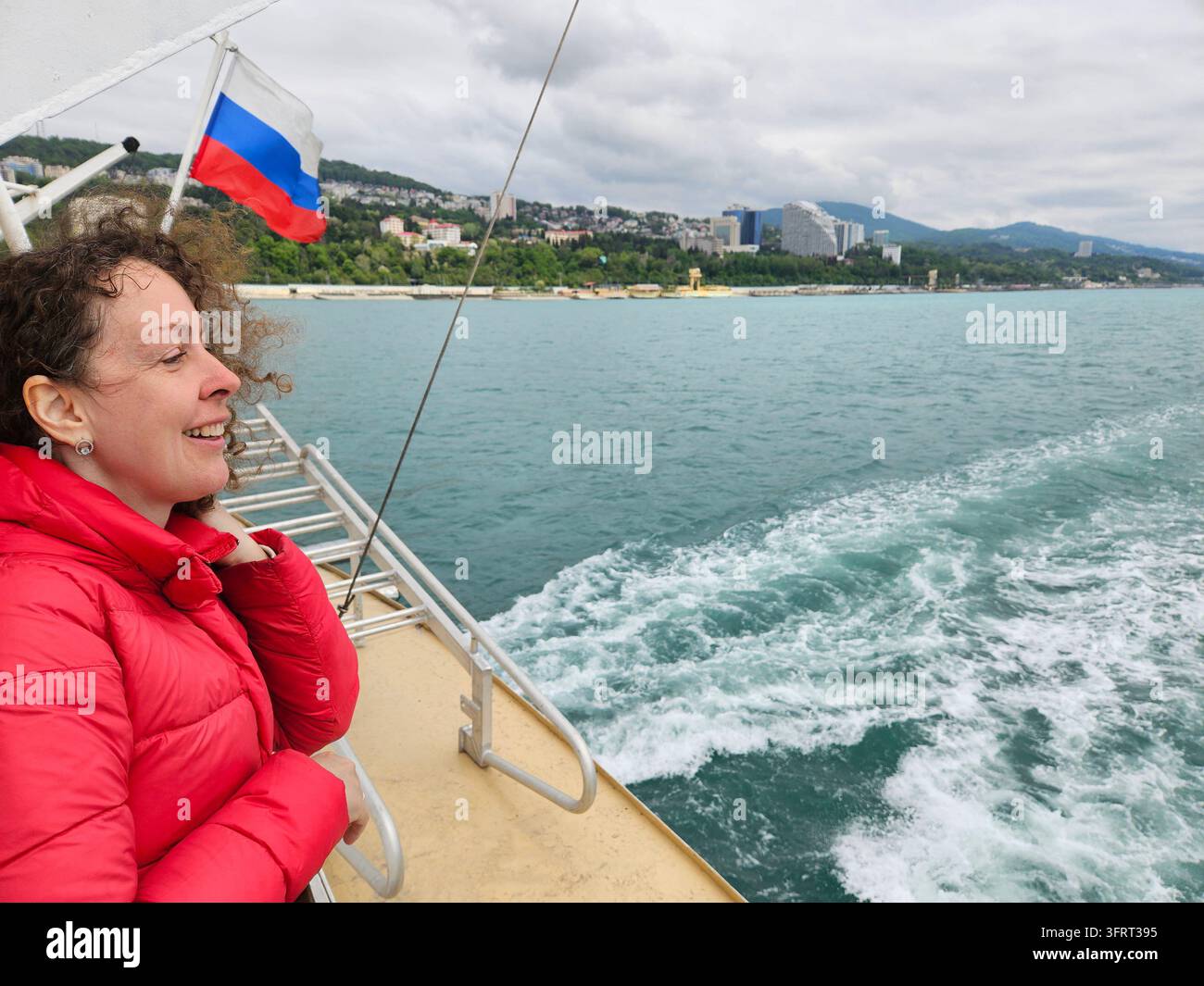 Frau in einer roten Jacke steht auf dem Heck eines Katamarans. Fahne über ihrer Hand Stockfoto