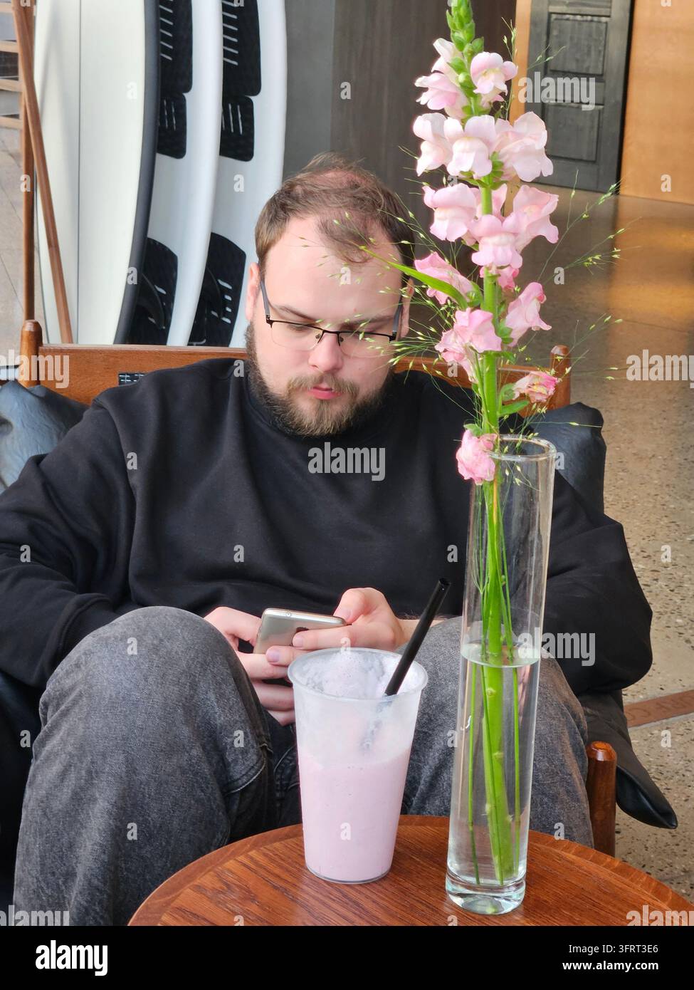 Mann sitzt auf einem Stuhl mit einer rosa Blume und einem Cocktail vor ihm. Er schaut auf sein Handy in der Nähe des Wakeboardings Stockfoto