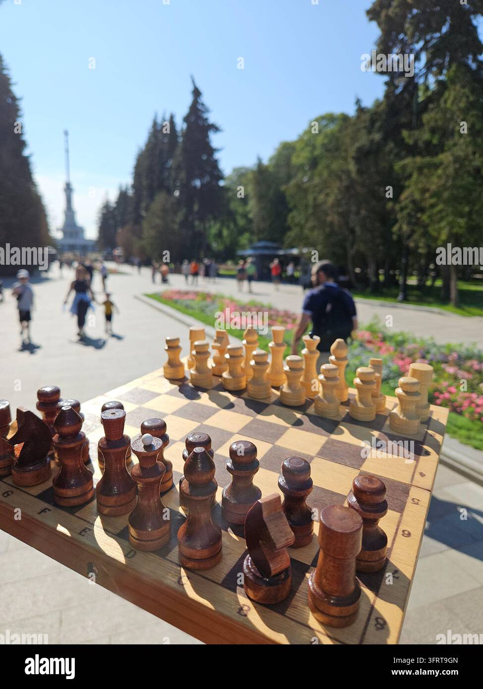 Ein hölzernes Schachbrett mit schwarzen und weißen Figuren wird von jemandem hochgehalten. Das Brett steht auf einem Bürgersteig vor einem Flussuferbahnhofspark. Die Szene ist friedlich und ruhig Stockfoto