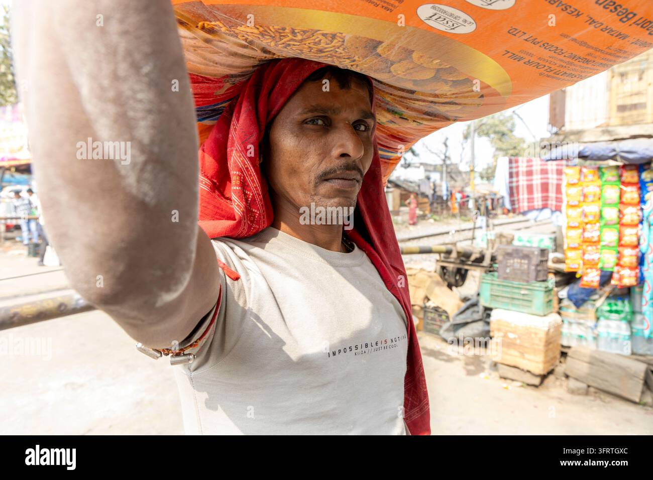 Ein Arbeiter hält einen schweren Sack auf einem roten Stoffpolster mit seinem Arm, der sich in Richtung Kamera erstreckt, während Imbissstände und Gleise hinter ihm stehen, West Bengal, Stockfoto