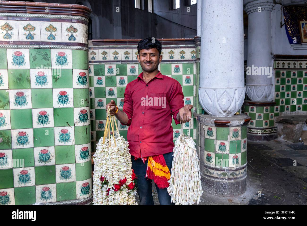 Stehender Verkäufer zeigt lange Stränge weißer Blumen mit einigen roten Blüten neben einer weißen Säule und einer karierten grün-weißen Fliesenwand auf dem Markt Stockfoto