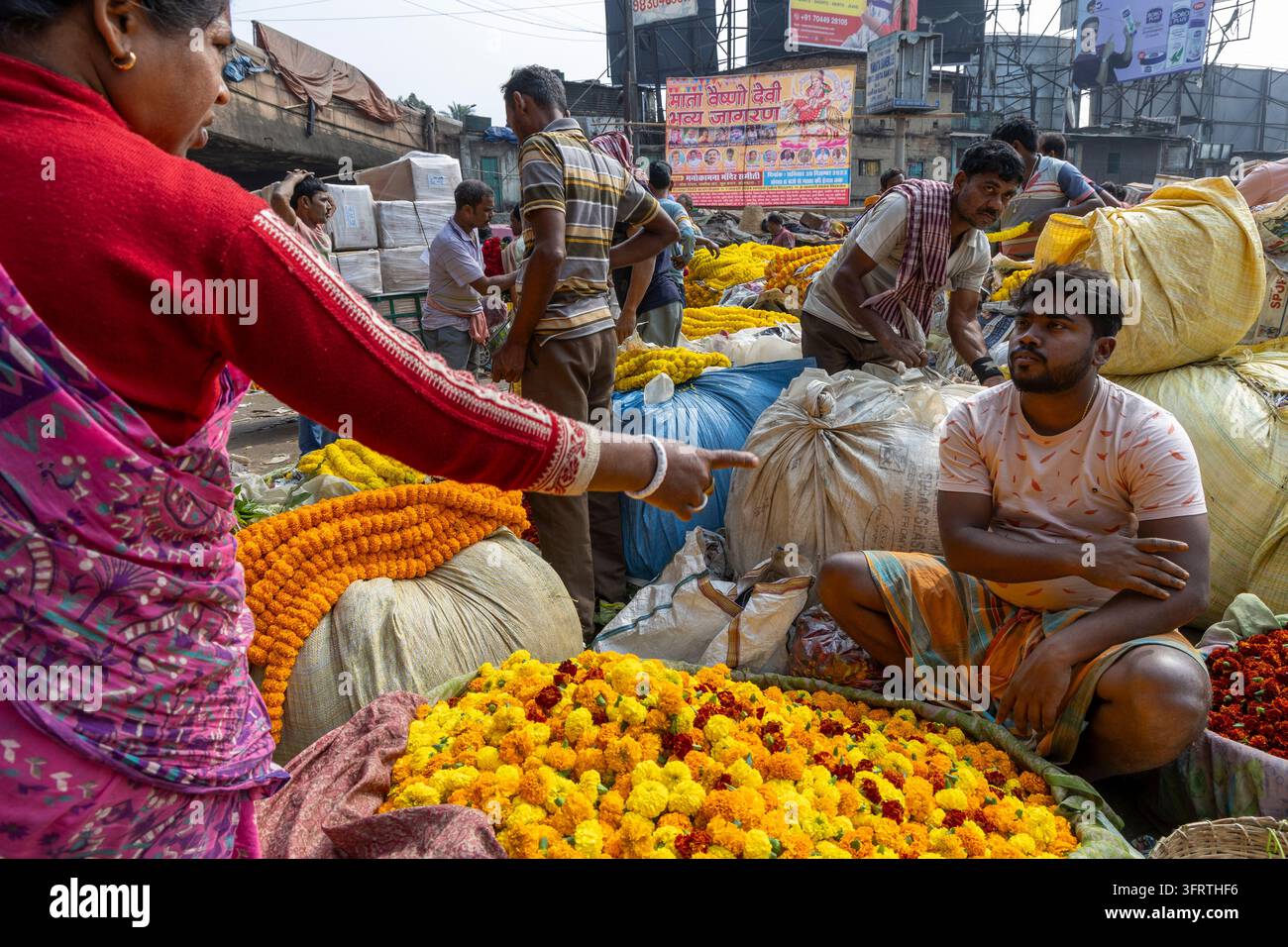 Kunde in einem rosa Sari deutet auf einen jungen Mann, der im Schneidersitz mit verschränkten Armen in der Nähe eines Korbs mit orangefarbenen und gelben Blumen am Blumenmarkt sitzt Stockfoto
