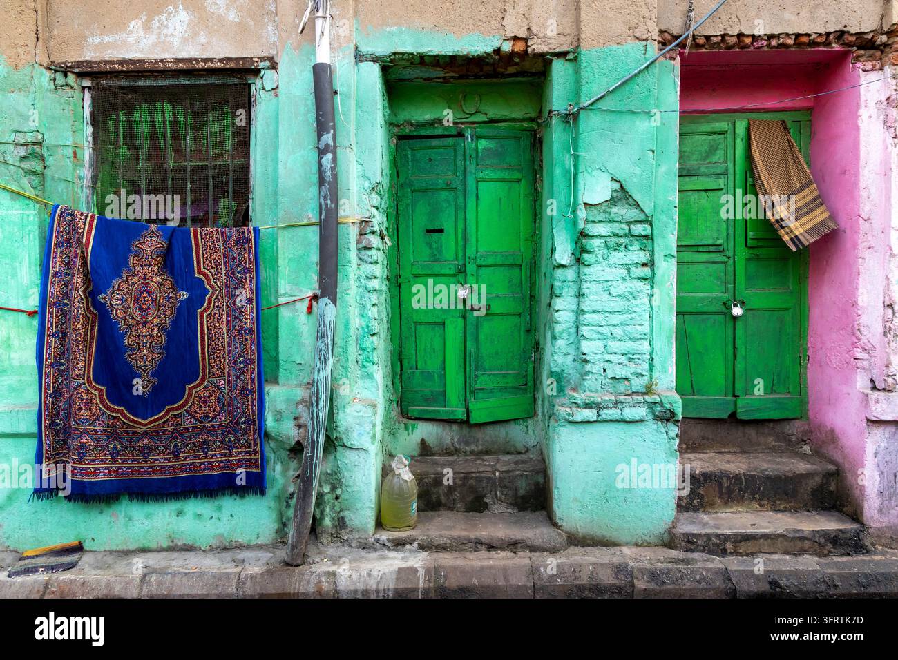 Ein blauer kunstvoller Teppich hängt unter einem vergitterten Fenster neben zwei geschlossenen grünen Holztüren in einer minzgrünen und rosa Wand mit bröckelndem Putz, West Bengal, Stockfoto