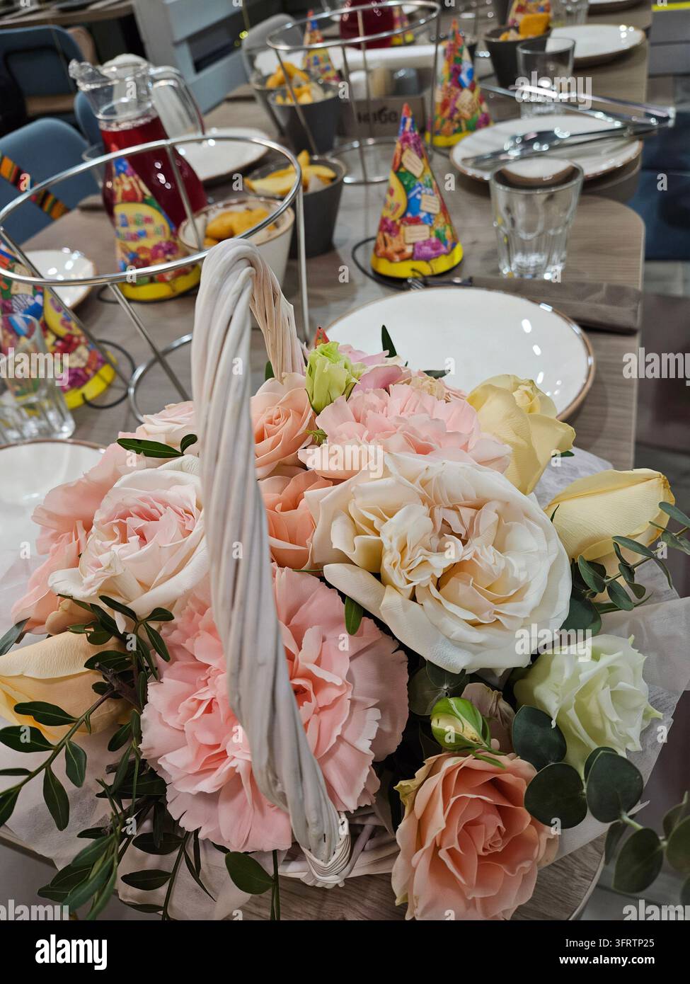Pastellrosen in einem gewebten Korb auf einem festlichen Tisch. Weiche rosa und cremefarbene Blumen mit Grünzeug. Tisch gedeckt für eine Feier mit bunten Partyhüten. Gläser, Teller und Getränke in der Nähe Stockfoto
