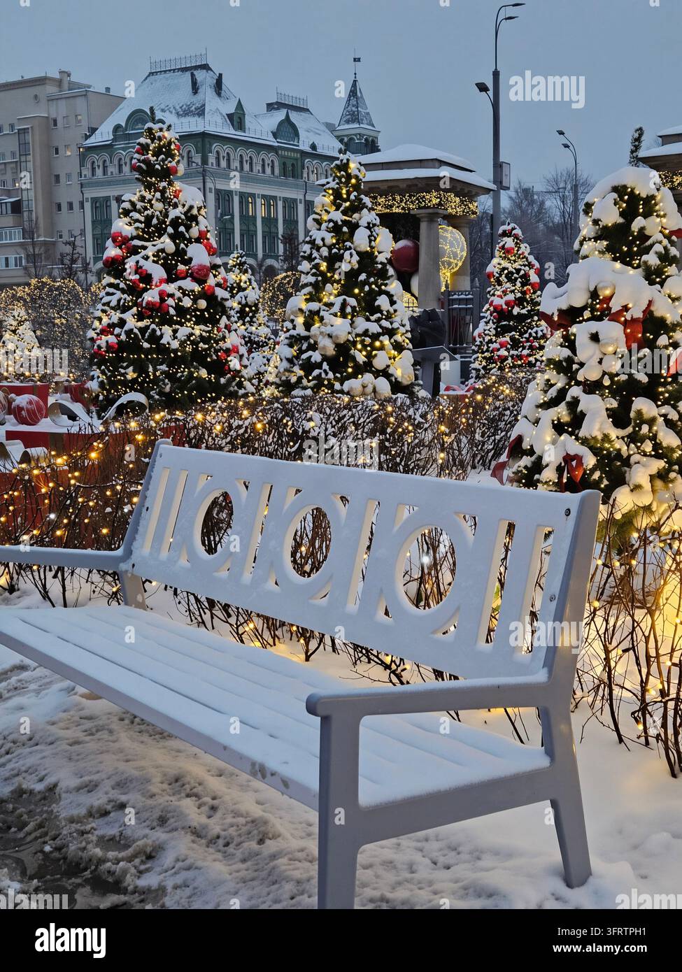Schnee bedeckt eine Bank neben leuchtenden, geschmückten Weihnachtsbäumen. Warme Lichter funkeln hinter den festlichen, schneebedeckten Bäumen. Der urbane Hintergrund zeigt schneebedeckte Gebäude und Straßenlaternen Stockfoto