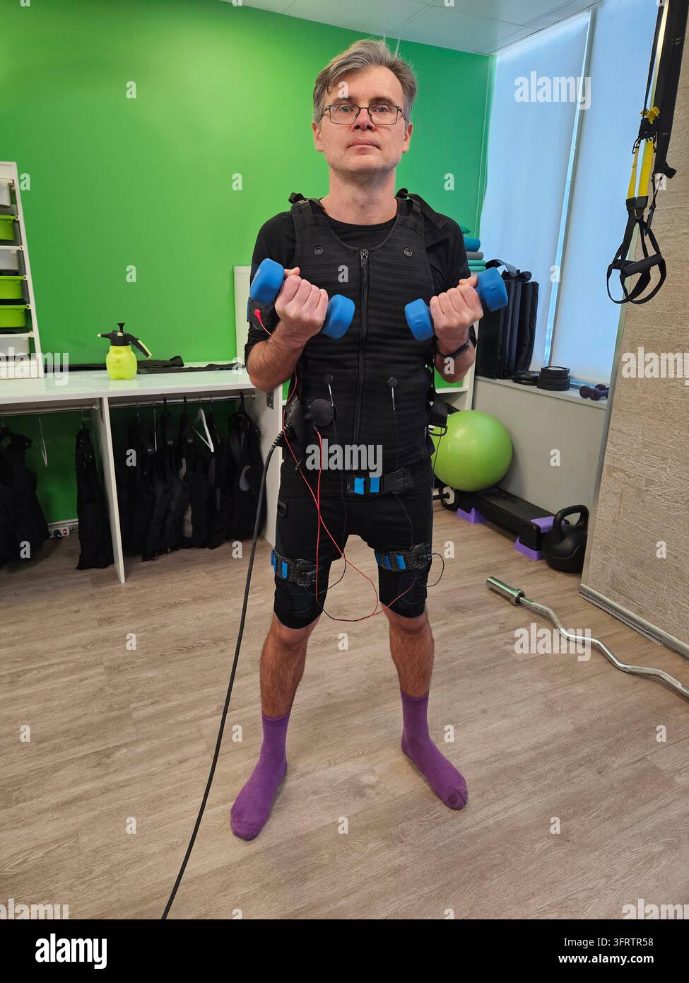 Mann trainiert mit Widerstandsbändern und Hanteln. Er trägt einen Activity-Tracking-Anzug mit Sensoren. Grüne Wand und Fitnessstudio-Ausrüstung umgeben ihn, Elektromuskelstimulation Stockfoto