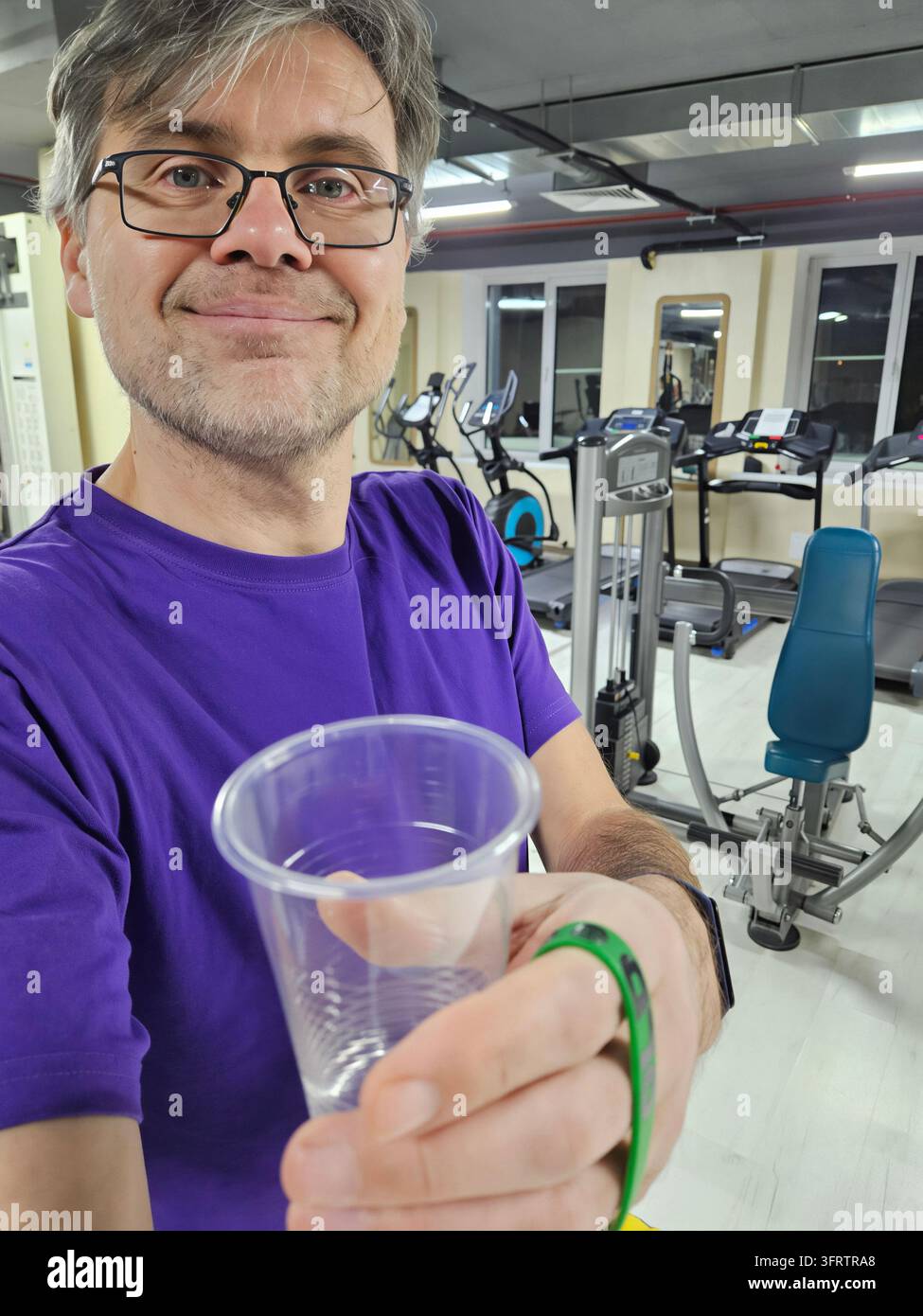 Mann hält einen klaren Plastikbecher in einer Fitnessstudio-Umgebung. Trägt ein lila Hemd und eine Brille. Fitnessstudio-Ausrüstung ist im Hintergrund sichtbar. Der Mann lächelt sanft in die Kamera. Die Hand zeigt auch ein grünes Armband Stockfoto