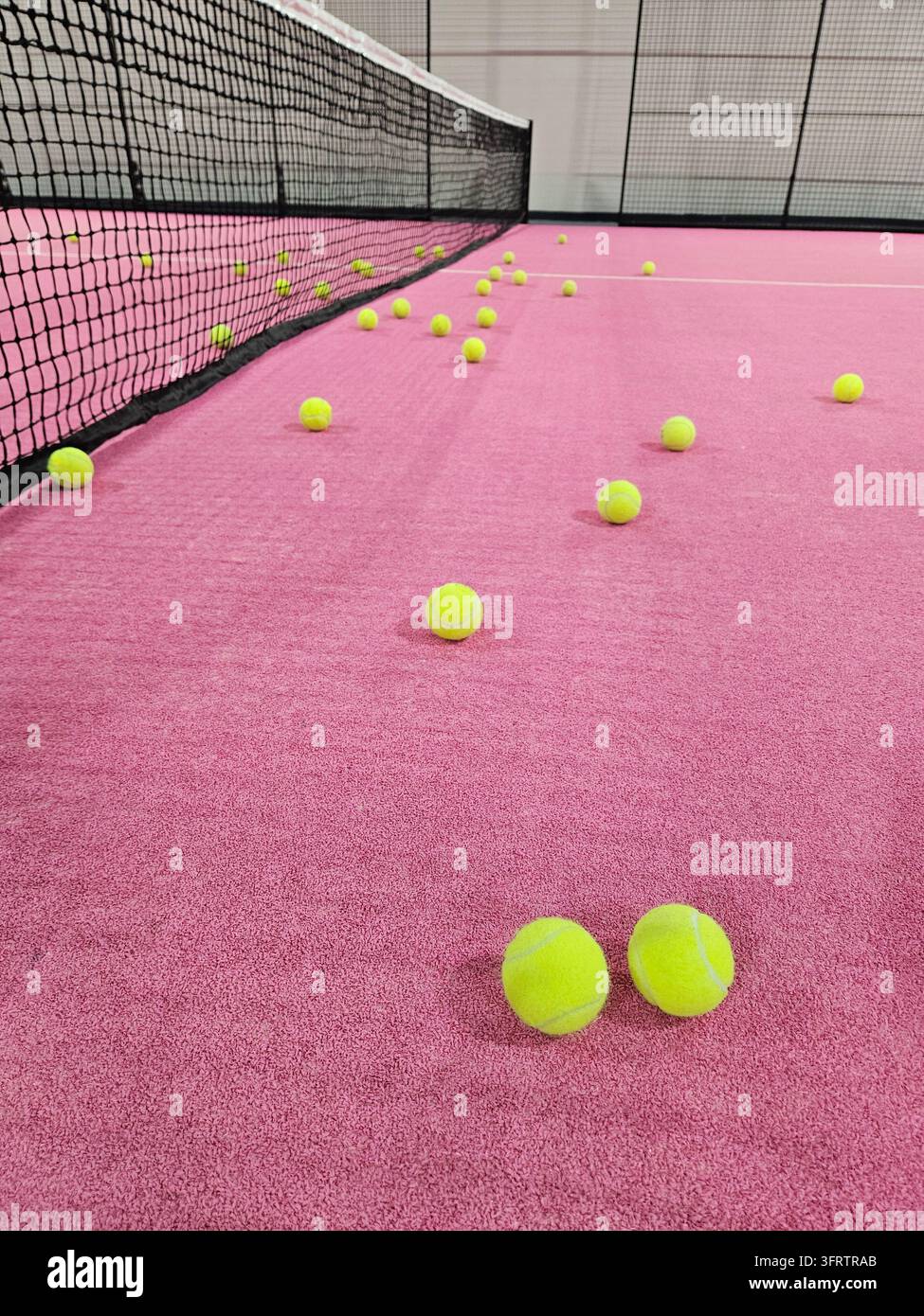 Rosa Tenniscourt mit verstreuten gelben Bällen. Schwarze Netzlinien säumen den Courtrand. Bälle ruhen auf einer strukturierten, lebendigen Oberfläche. Perfekt für Sport, Training oder athletische Bilddarstellung Stockfoto