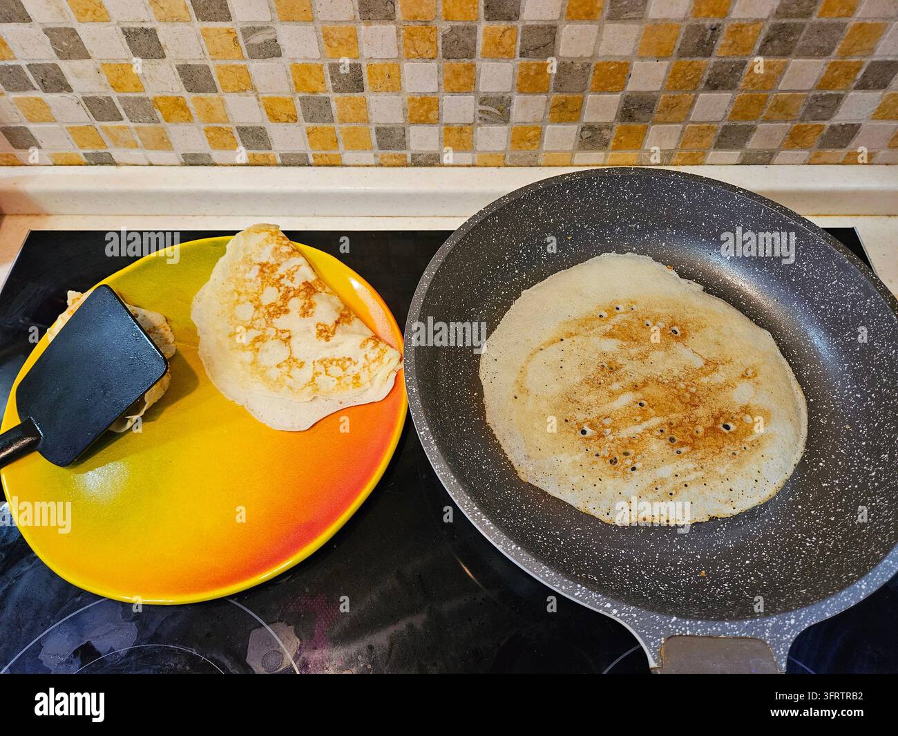 Knuspriger Pfannkuchen brät in einer gesprenkelten Pfanne auf dem Herd. Der zweite Pfannkuchen liegt auf einem bunten Teller neben dem Pfannenwender. Im Hintergrund sind gemusterte Küchenfliesen zu sehen. Das Essen ist frisch zubereitet und servierfertig Stockfoto