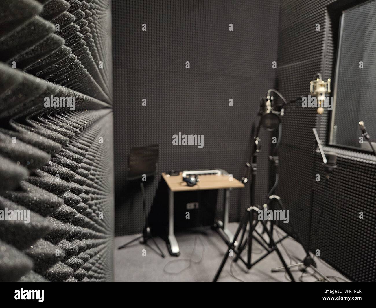 Professionelles Aufnahmestudio mit Akustikpanelen und Mikrofonen. Geräteständer stehen in der Nähe eines Schreibtisches mit einer Kaffeetasse. Schwarz verkleideter Raum für Audioproduktion oder Voiceover Stockfoto
