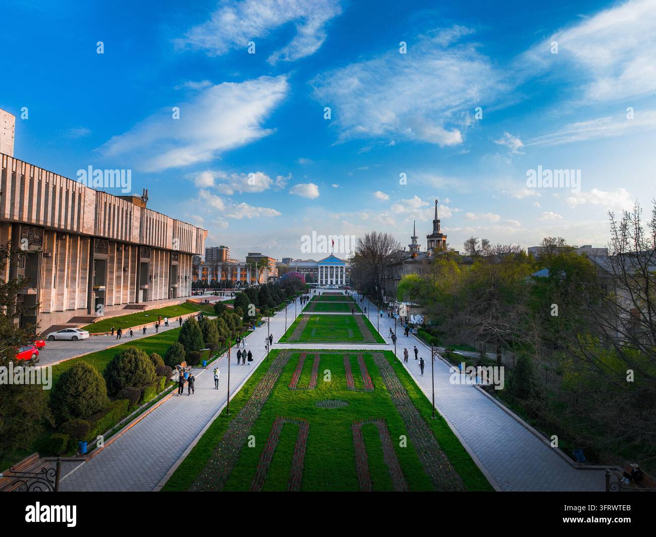 Befestigte Promenade, gepflegte Rasenflächen und Bäume zwischen öffentlichen Gebäuden in Bischkek unter blauem Himmel Stockfoto