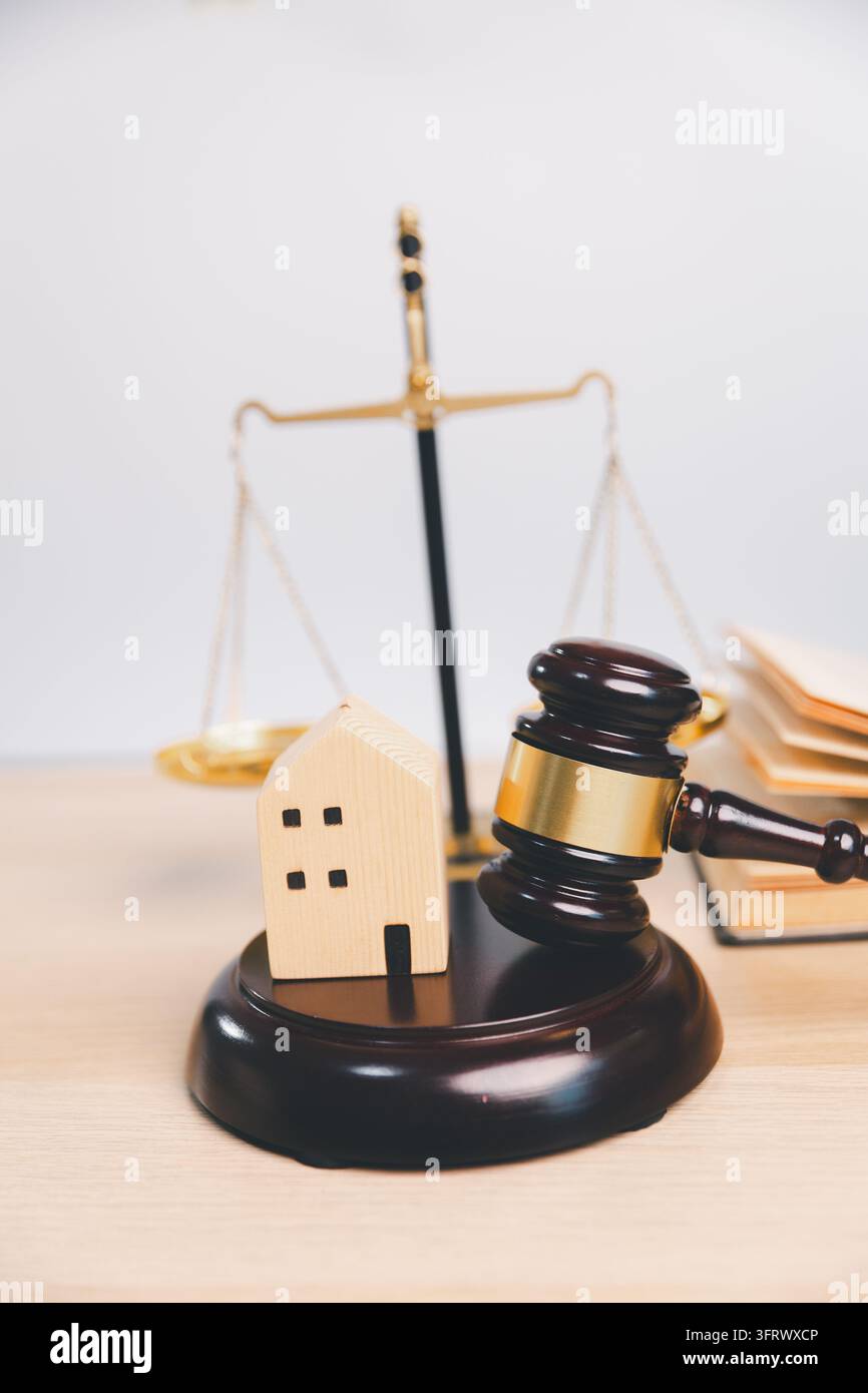 Ein kleines Holzhaus und ein Hammer sitzen auf einem dunklen Sockel vor einer Waage der Gerechtigkeit und einem offenen Buch, das ein Grundstücksrechtskonzept mit großzügigem Li illustriert Stockfoto