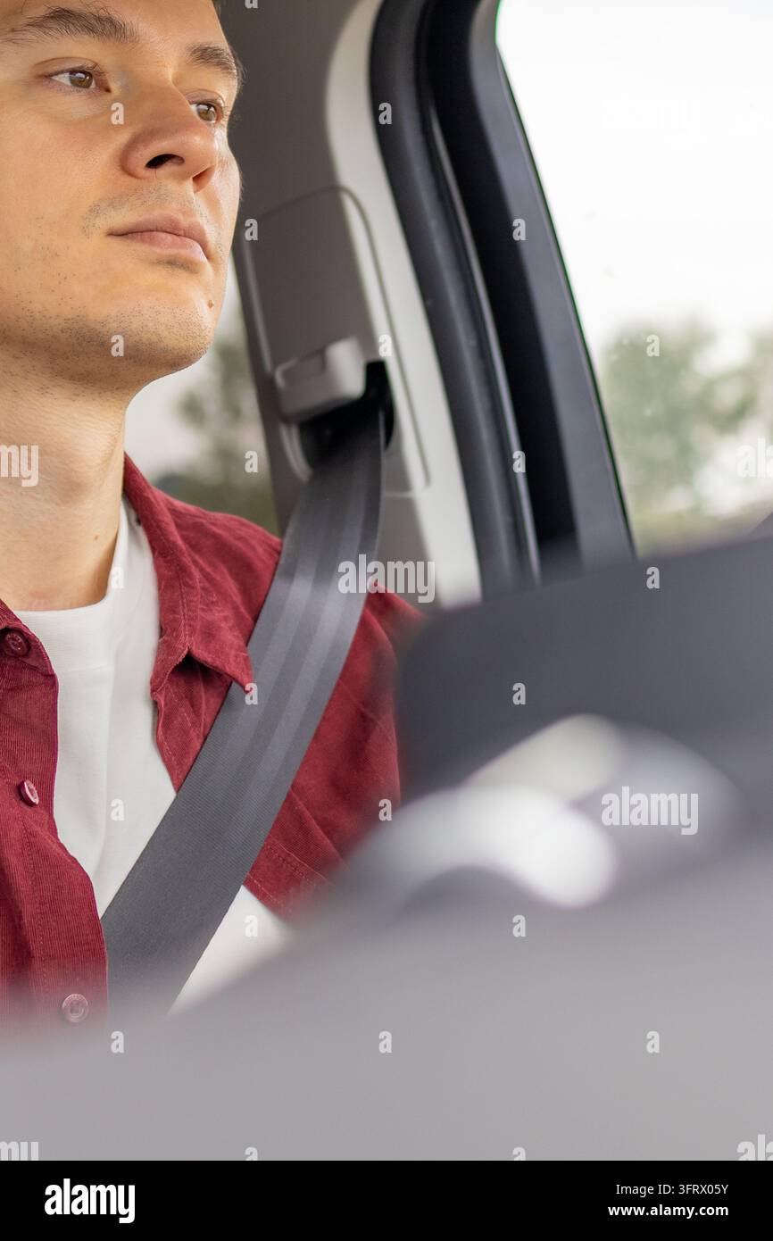 Nahaufnahme eines ernsten jungen Mannes in einem roten Cordhemd mit angelegtem Sicherheitsgurt, der während der Autofahrt nach vorne auf die Straße schaut. Konzept eines konzentrierten d Stockfoto