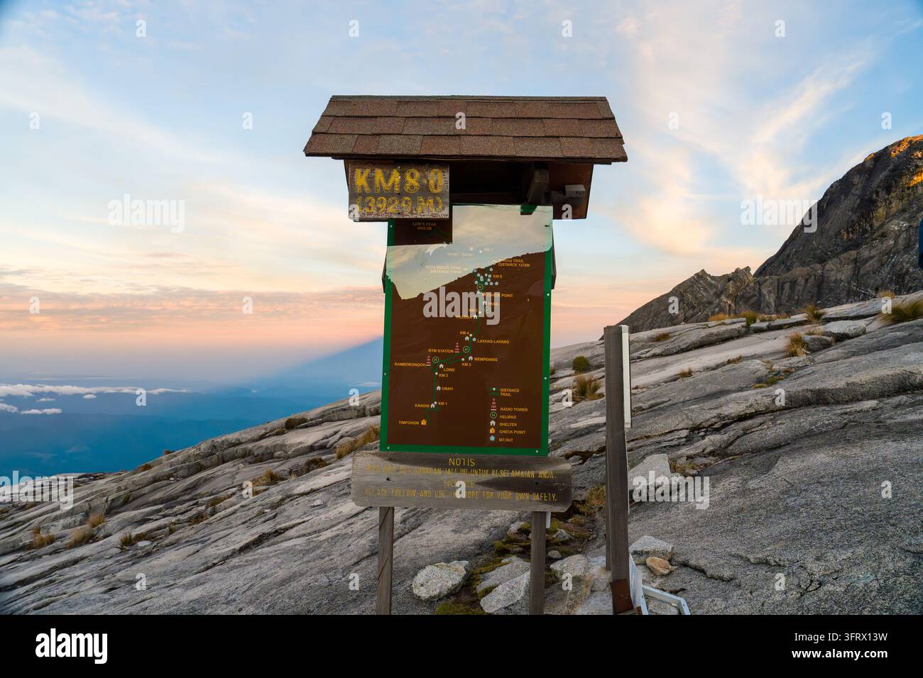 Berggipfelwegweiser bei Sonnenaufgang. Felsen unter sanftem goldenem Licht. Wegweiser mit Karte und Textdetails. Ferne Hügel und Täler darunter. Cle Stockfoto