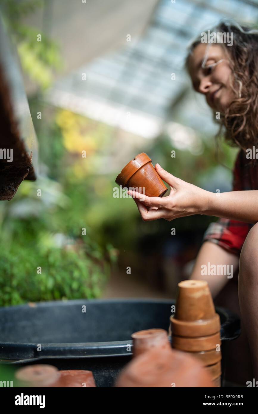 Lächelnde Frau arbeitet in botanischem Garten, sitzt und reinigt Keramikblumentöpfe in einem Wassereimer. Stockfoto
