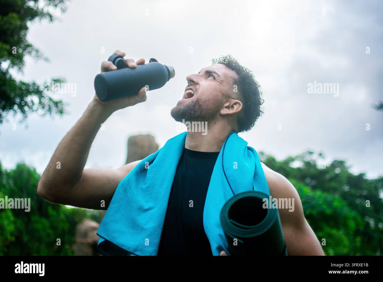 Niedrigwinkelaufnahme eines sportlichen Mannes, der Wasser aus einer wiederverwendbaren Sportflasche trinkt, während er eine gerollte Yogamatte trägt und ein blaues Handtuch um den Nacken trägt Stockfoto
