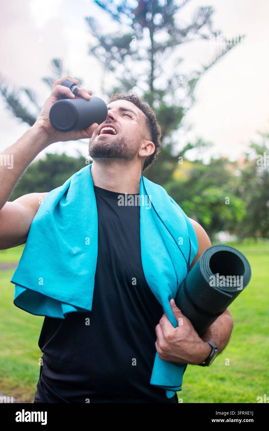 Niedrigwinkelaufnahme eines sportlichen Mannes, der Wasser aus einer wiederverwendbaren Sportflasche trinkt, während er eine gerollte Yogamatte trägt und ein blaues Handtuch um den Nacken trägt Stockfoto