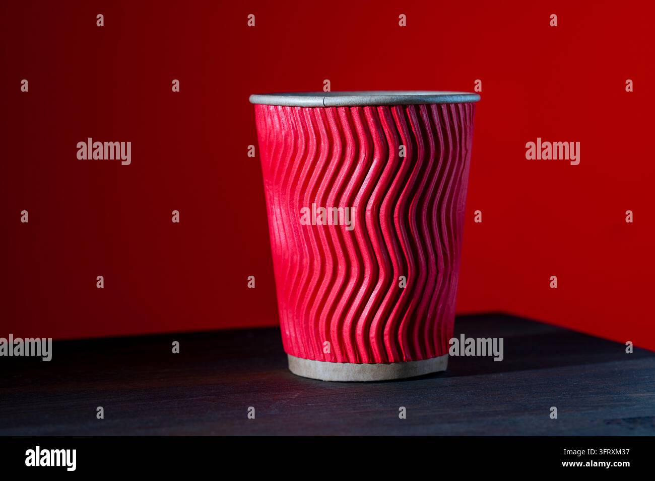 Roter wellenförmiger Pappkaffeebecher auf dunklem Holztisch. Nahaufnahme eines roten, gerippten Einweg-Pappbechers für Heißgetränke. Zum Mitnehmen-Kaffeebecher auf dunklem Holz de Stockfoto