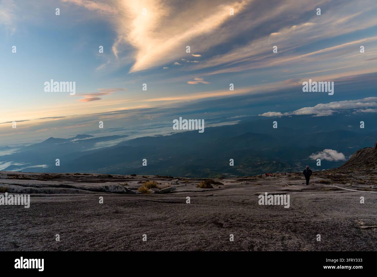 Einsamer Wanderer steht auf einem schroffen Berggipfel. Ferne Wolken ziehen über den weiten Horizont. Sonnenlicht taucht den Himmel in sanfte goldene Farbtöne. Das Gelände ist felsig Stockfoto