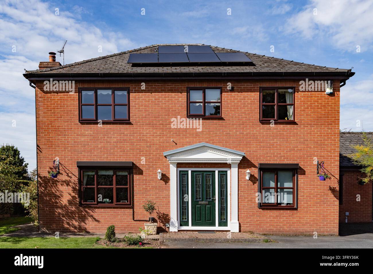 Vorderansicht eines symmetrischen freistehenden Hauses aus rotem Ziegelstein mit Solarpanelen auf einem schrägen grauen Ziegeldach. Stockfoto