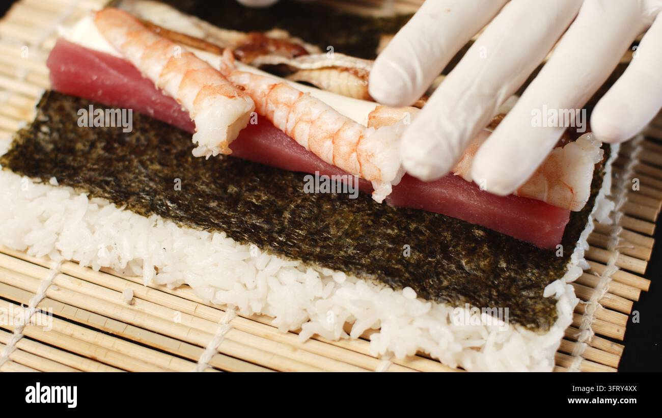Chef-Hände in Handschuhen bereiten eine frische Sushirolle mit Thunfisch, Garnelen, Reis und Nori auf einer Bambusmatte zu Stockfoto