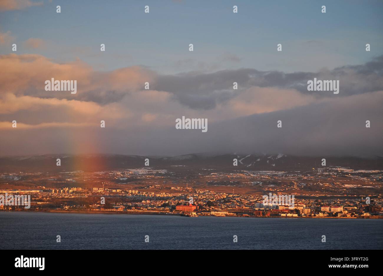Panoramatische Küstenstadt bei Sonnenuntergang mit einem schwachen Regenbogen, Bergen und dramatischen Wolken Stockfoto