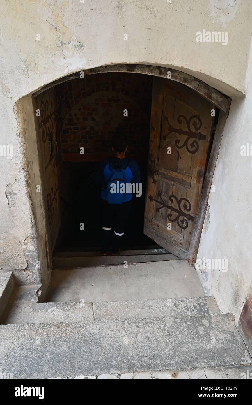 Eine Touristin mit einem Rucksack, die durch eine offene Holztür in einen dunklen Burgenkellerflur eintritt. Stockfoto