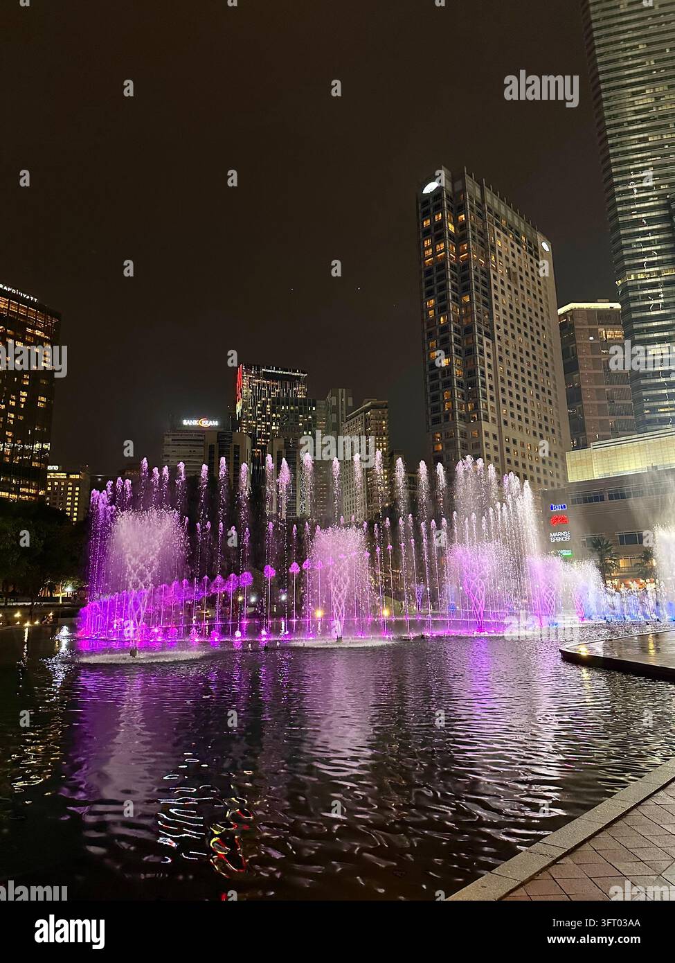 Kuala Lumpur, Malaysia – 26. November 2022: Rosa beleuchtete Fontänen spiegeln sich unter den Hochhäusern von Kuala Lumpur in einer nächtlichen städtischen Szenerie - Smartphone-aufgenommenes Stockfoto
