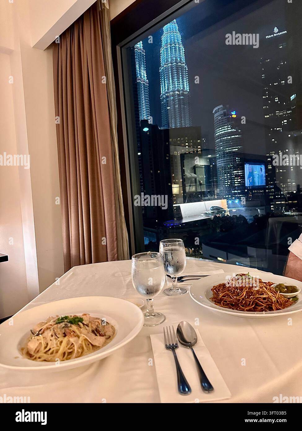 Pasta-Gerichte stehen neben einem Fenster mit Blick auf die beleuchteten Petronas-Zwillingstürme während des Abendessens. - Smartphone-aufgenommenes Stockfoto