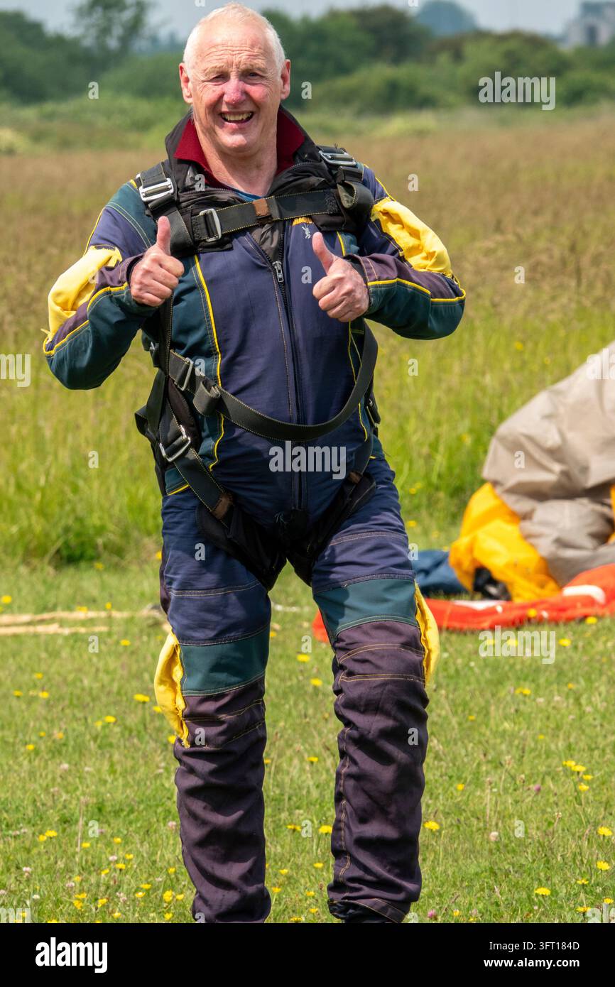 Ein glücklicher Senior lächelt und zeigt einen doppelten Daumen nach oben, nachdem er erfolgreich einen Fallschirmsprung auf einem grasbewachsenen Flugplatz an einem hellen Sommertag gelandet hat. Stockfoto