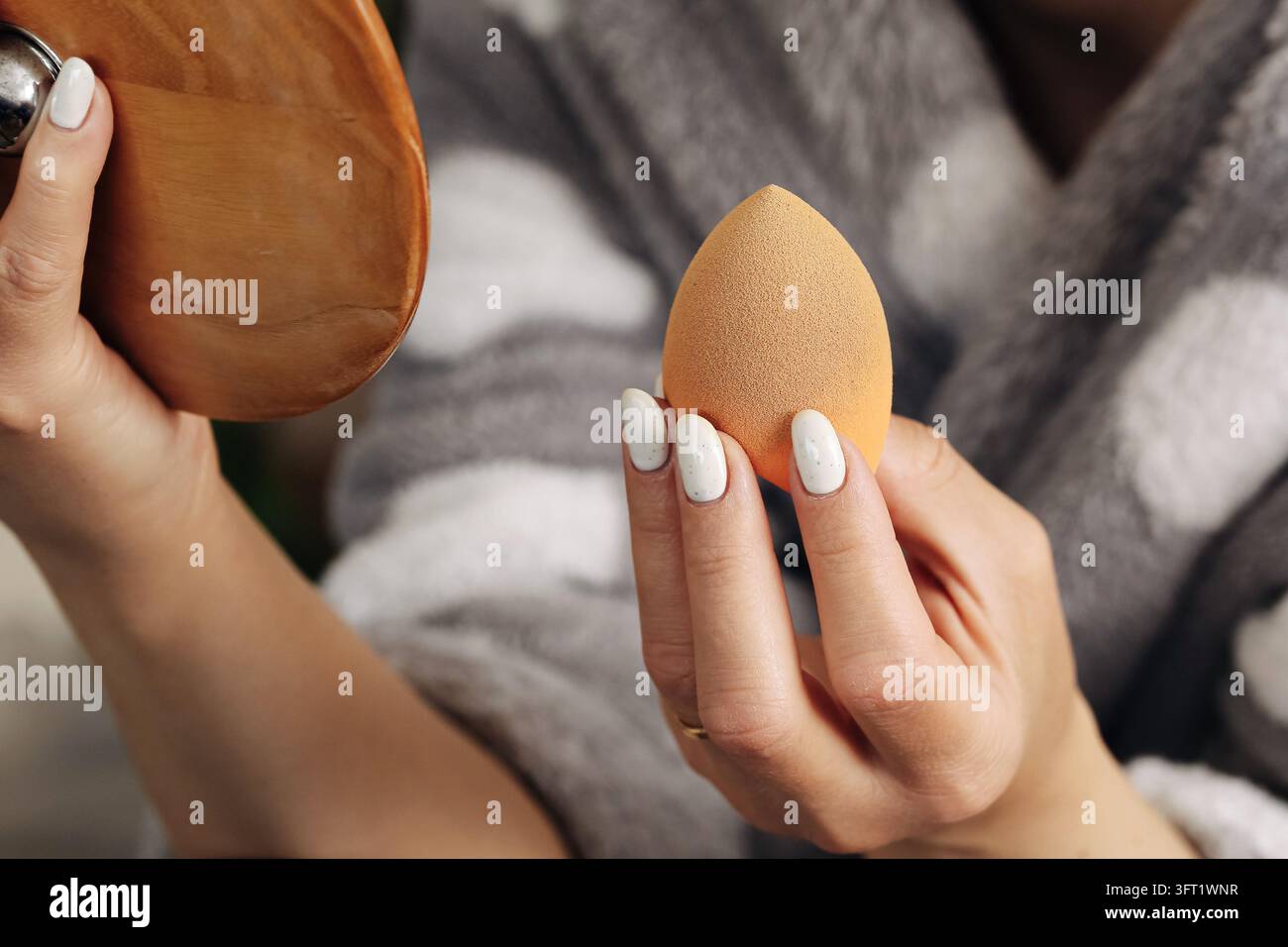 Frau hält Make-up-Schwamm und Kompaktspiegel während der Schönheitsroutine. Kosmetik-Hintergrund für Grundierung, Gesichts-Make-up und persönliche Pflege Stockfoto