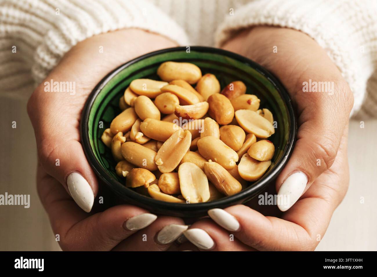 Frau hält eine grüne Schüssel mit geschälten Erdnüssen in beiden Händen. Gesunder Snack-Hintergrund für gesalzene Nüsse, eiweißreiche Lebensmittel und lockeres Essen. Stockfoto