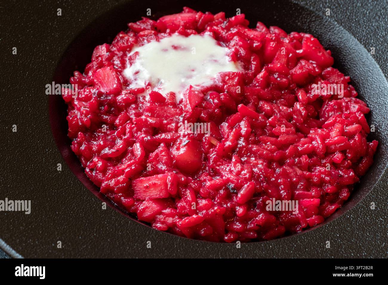 Hochwinkelsicht von rubinrotem Rhabarber-Risotto mit Apfelwürfeln und Taleggio-Fondue in einer schwarzen Schüssel auf einer dunklen Arbeitsplatte, weiches natürliches Licht Stockfoto