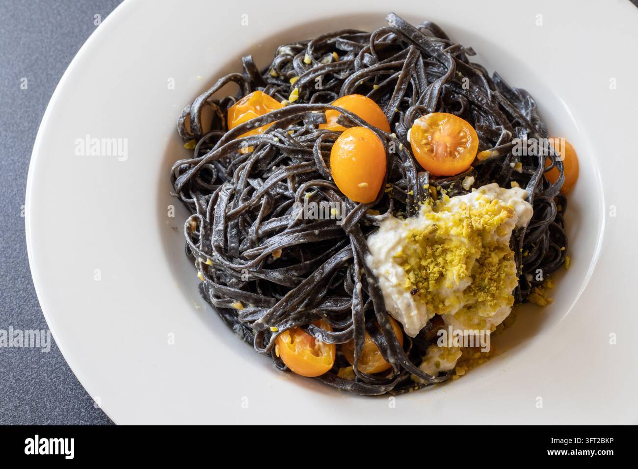 Hohe Blickwinkelaufnahme von schwarzer Tintenfisch-Tintenlinguine mit Burrata und Pistazien in einer weißen Schüssel auf einer dunklen Arbeitsplatte, mit gelben Kirschtomaten Stockfoto